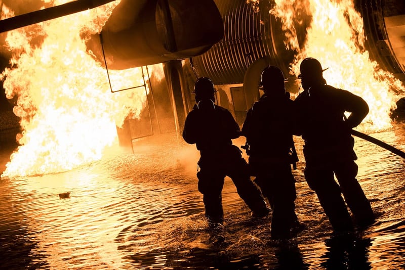 Three firefighters silhouetted against flames at a night fire, standing in water with a hose line.