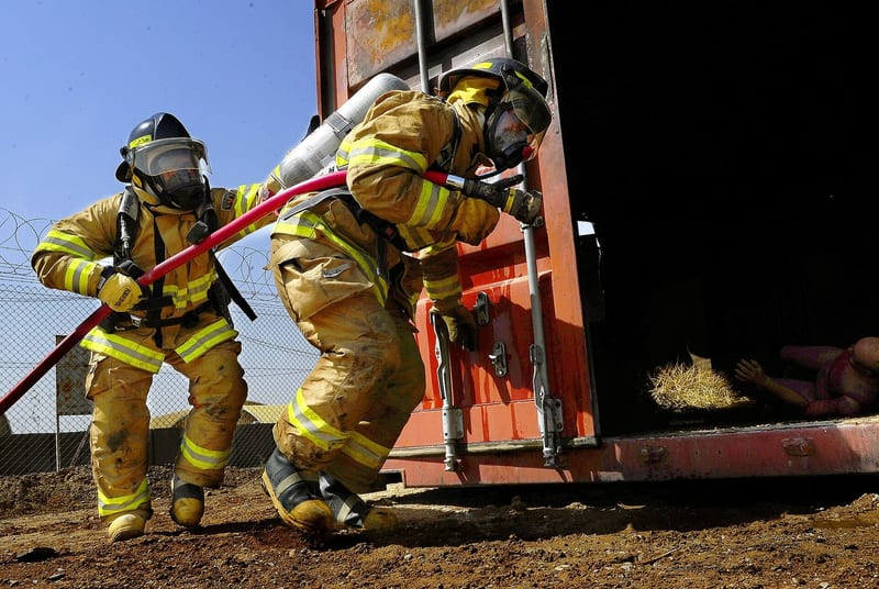 Two firefighters in full gear and air packs advance a charged hose line toward a training container.